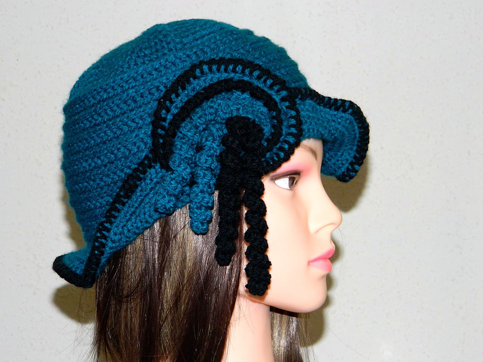 Handgehäkelte Cloche in Petrol mit schwarzer Schnecke und Korkenzieher-Locken auf einem Modisten-Kopf